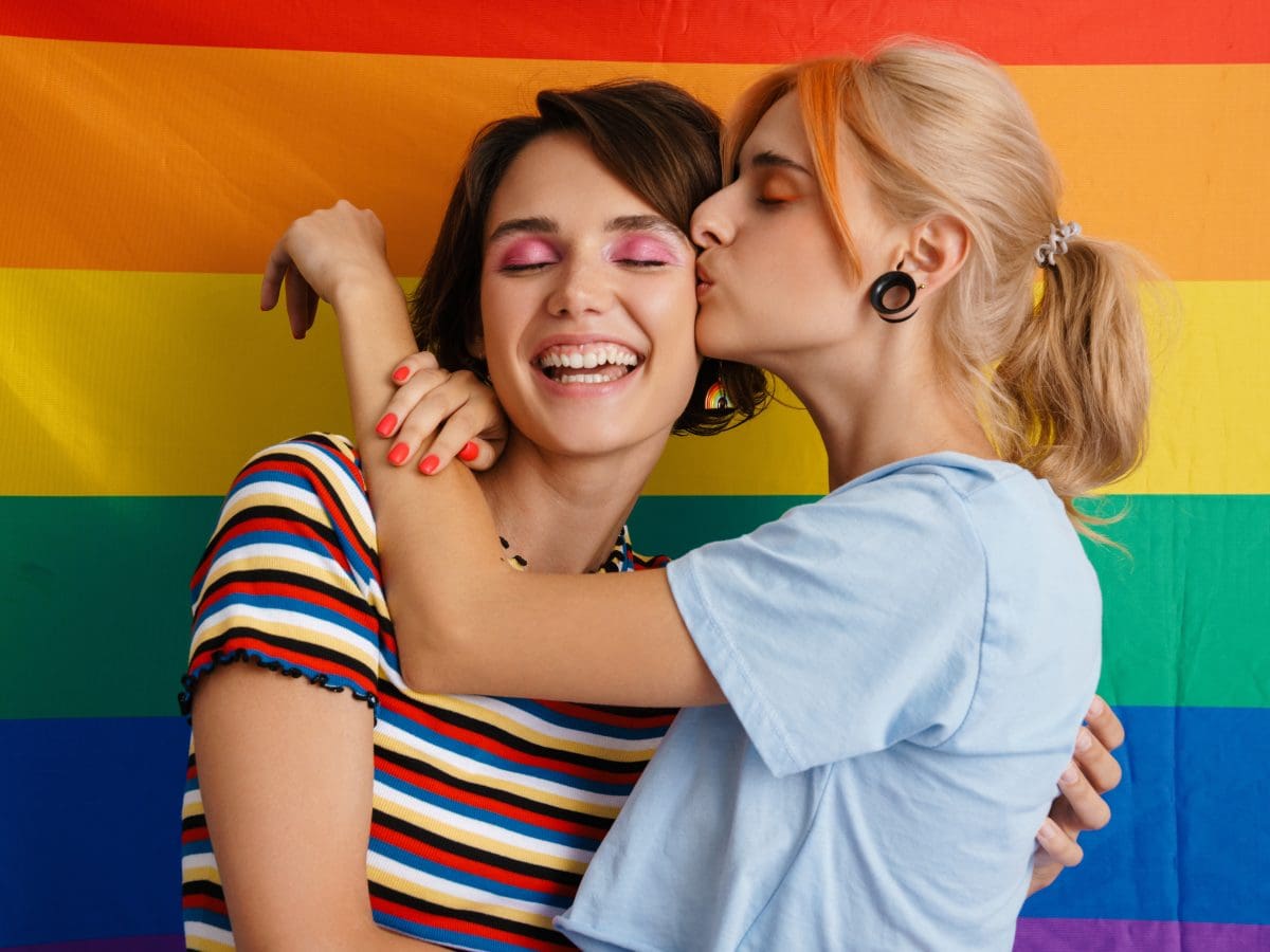 Two young women embracing in front of a rainbow flag